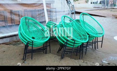 Korbgrüne Plastikstühle mit schwarzen Beinen in einem Café an einem Sandstrand nach einem Sturm. Die Möbel sind übereinander gestapelt, da das Strandcafe geschlossen ist. Ende der Strandsaison im Hotel. Stockfoto