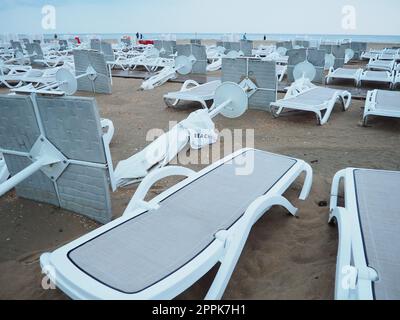 Ein Strandschirm liegt nach schlechtem Wetter auf einem Sandstrand gefaltet. Sturm auf See. Ende der Strandsaison durch Taifun und Zyklon. Liegestühle, Sonnenschirme und Hotelmöbel. Windgedrehte Strukturen Stockfoto