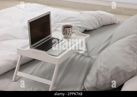 Weißer Tablett-Tisch mit Laptop und einer Tasse Getränk auf dem Bett Stockfoto