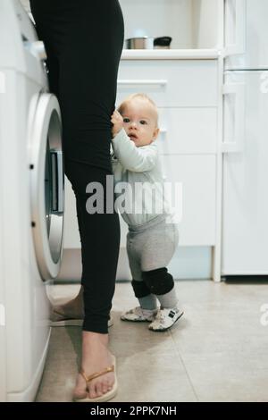 Ein kleines kleines Kind, das sich zwischen den Beinen der Mutter versteckt und ihre Aufmerksamkeit während des Multitaskings verlangt, während es versucht, Hausaufgaben in der Küche zu Hause zu erledigen. Mutter im Mutterschaftsurlaub. Stockfoto