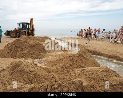 Anapa, Russland, 19. August 2021 Ein Traktor gräbt einen Graben an einem Sandstrand, um verschüttetes Abwasser nach langen Regenfällen abzuleiten. Leute, die Touristen sehen sich Erdbauarbeiten an. Abstieg des Wassers ins Meer Stockfoto