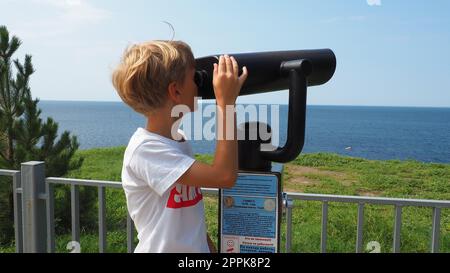 Anapa, Russland, 23. August 2021 Ein Junge schaut durch ein Teleskop. Ein 9-jähriges kaukasisches Kind hält ein großes marines Teleskop mit seinen Händen. Die Straße der Resortstadt und Wandertouristen Stockfoto