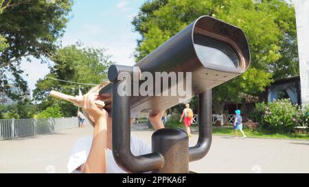 Anapa, Russland, 23. August 2021 Ein Junge schaut durch ein Teleskop. Ein 9-jähriges kaukasisches Kind hält ein großes marines Teleskop mit seinen Händen. Die Straße der Resortstadt und Wandertouristen Stockfoto