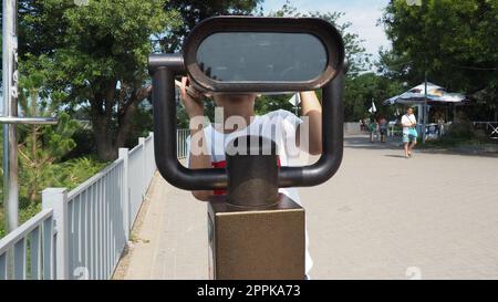 Anapa, Russland, 23. August 2021 Ein Junge schaut durch ein Teleskop. Ein 9-jähriges kaukasisches Kind hält ein großes marines Teleskop mit seinen Händen. Die Straße der Resortstadt und Wandertouristen Stockfoto