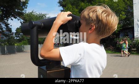 Anapa, Russland, 23. August 2021 Ein Junge schaut durch ein Teleskop. Ein 9-jähriges kaukasisches Kind hält ein großes marines Teleskop mit seinen Händen. Die Straße der Resortstadt und Wandertouristen Stockfoto