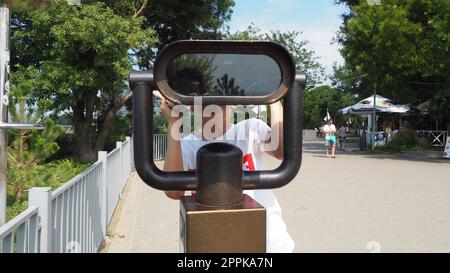 Anapa, Russland, 23. August 2021 Ein Junge schaut durch ein Teleskop. Ein 9-jähriges kaukasisches Kind hält ein großes marines Teleskop mit seinen Händen. Die Straße der Resortstadt und Wandertouristen Stockfoto