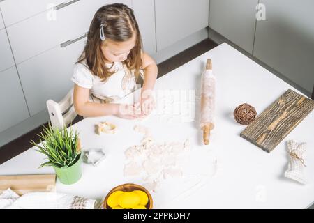 Porträt einer kleinen, ernsten Küchenchefin, die hilft und osterkekse macht. Formen Sie Teigmehl, Backwaren, Becher, Kekse. Stockfoto