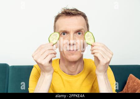 Glücklicher erwachsener Mann, der Augenflecken aus frischen Gurkenscheiben in den Händen hält. Feuchtigkeitsmaske für dunkle Augenkreise. Stockfoto