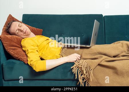 Müder Mann schläft auf dem Sofa mit offenem Laptop im Wohnzimmer. Überarbeitende Person, die sich auf dem Kissen unter der Decke zurücklehnt. Stockfoto