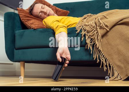 Ein müder Mann schläft auf dem Sofa mit einem Smartphone in der Hand im Zimmer. Überarbeitende Person, die sich auf dem Kissen unter der Decke zurücklehnt. Stockfoto