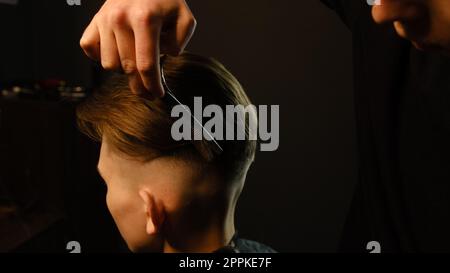 Der Friseur verwendet eine dünnere Schere und einen Metallkamm auf braunem, welligem Haar eines jungen Mannes. Friseurservice in einem modernen Friseursalon in einem dunklen Licht mit warmem Licht Stockfoto