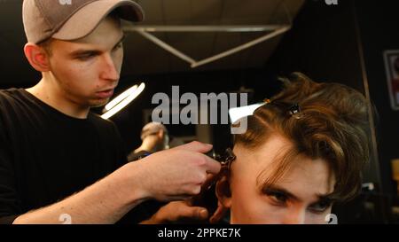 Aufnahme eines gutaussehenden Friseurs, der seinem Klienten mit dem Trimmer die Haare schneidet. Friseurservice in einem modernen Friseursalon mit dunklem Licht und warmem Seitenblick Stockfoto