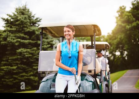 Es ist fast Zeit, zurück zum Clubhaus zu gehen. Eine attraktive junge Frau, die sich auf einem Golfplatz gegen einen Golfwagen lehnt. Stockfoto