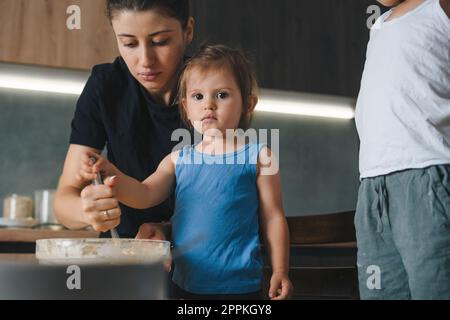 Süßes kleines Mädchen und ihre wunderschöne Mutter, die Eier wackelt, während sie zu Hause backt und in die Kamera schaut. Hobby-Kochbegriff für den Haushalt. Stockfoto