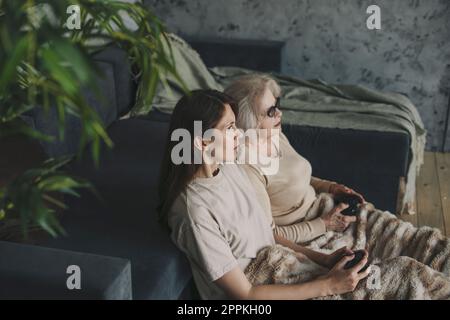 Erwachsene Frau und ältere Mutter, die Spaß haben, Zeit miteinander verbringen, Videospiele mit Gamepads zu Hause spielen. Aktive moderne ältere Menschen. Stockfoto