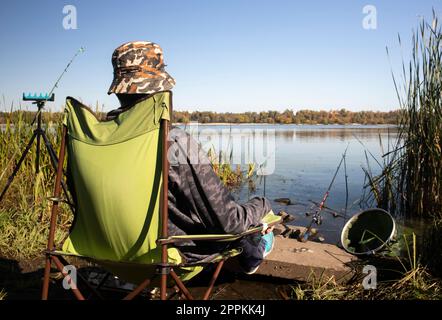 Ein junger, nicht wiedererkennbarer Fischer fängt Fische auf einem Stuhl am Ufer eines Flusses oder Sees. Der Angler sitzt auf einem Stuhl mit Angelausrüstung. Ein perfekter Urlaub am Fluss mit einer Angelrute. Stockfoto