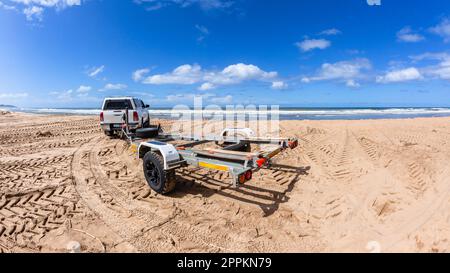 Leerer Bootsanhänger am Strand, am Ufer entlang, mit Bootsbesitzern, die in der Meereslandschaft fischen. Stockfoto