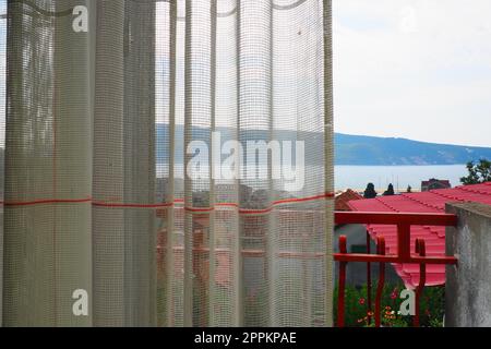 Weiße, grobe Baumwolle- oder Leinenvorhänge aus Gewebungen aus dünnen Fäden. Einfache, kompakte und kostengünstige Wohnungseinrichtung. Fensterdekoration im Zimmer. Roter Streifen. Blick vom Hotelzimmer auf das Meer. Stockfoto