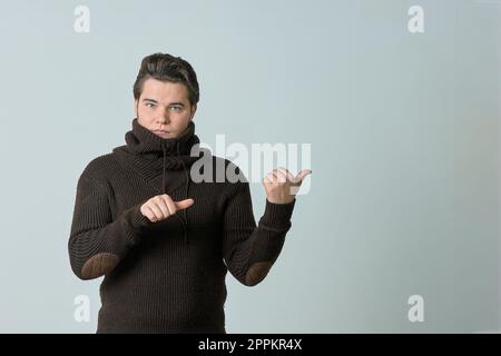 Ein Mann in einem braunen Pullover und einer leuchtenden Farbe zeigt auf etwas und perfekt, zeigt einen leeren Bereich mit seinem Daumen, zeigt alles auf einem hellen Hintergrund, Kopierbereich Stockfoto