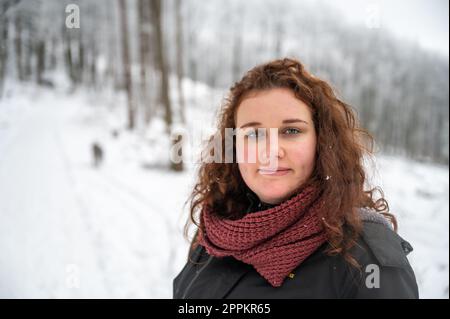 Eine junge Frau mit braunem lockigem Haar und warmer Kleidung lächelt und schaut in die Kamera, geht im Winter mit viel Schnee mit ihrem grauen akita inu Hund im Wald spazieren Stockfoto