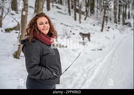 Eine junge Frau mit braunem lockigem Haar und warmer Kleidung lächelt und schaut in die Kamera, geht im Winter mit viel Schnee mit ihrem grauen akita inu Hund im Wald spazieren Stockfoto