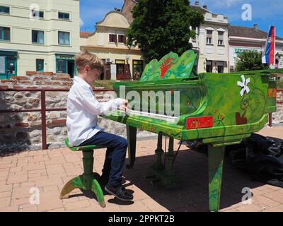 Sremska Mitrovica, Serbien, 05. Juni 2021. Ein Junge in einem weißen Hemd spielt auf der Straße Klavier. Das Kind ist 9 Jahre alt, weiß. Klavierfarben grün. Aufführung eines Musikstücks. Straßenkonzert Stockfoto