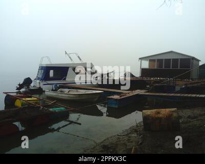 Belgrad, Serbien, 14. Januar 2020 Weißes Boot am Pier, Abfahrt für den Winter. Nebiges Wetter an einem wolkigen Januar-Tag. Reflexion im Fluss. Ein Haus zum Schutz vor dem Wetter Stockfoto