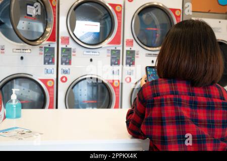 Rückansicht einer Frau, die mit einem Handy auf die Wäsche wartete, die in der Waschmaschine im Waschsalon gewaschen wurde. Eine Reihe industrieller Waschmaschinen im Münzwaschsalon. Stockfoto