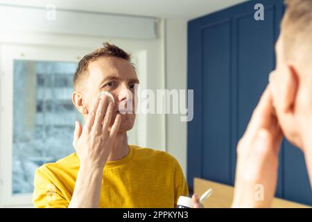 Ein erwachsener Mann trägt Gesichtscreme mit Wattepad auf und massiert dann mit den Händen. Männliches Porträt im Spiegel. Gesichtsreinigung. Stockfoto