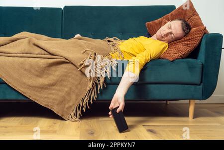 Ein müder Mann schläft auf dem Sofa mit einem Smartphone in der Hand im Zimmer. Überarbeitende Person, die sich auf dem Kissen unter der Decke zurücklehnt. Stockfoto