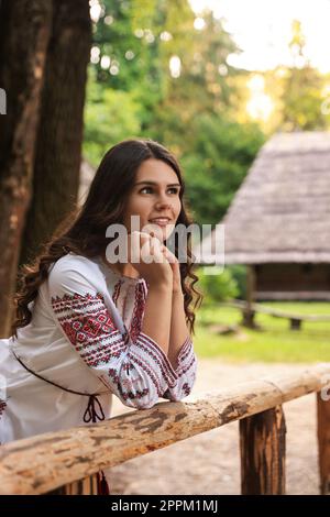 Schöne Frau, die ein besticktes Hemd neben einem Holzgeländer auf dem Land trägt. Ukrainische Nationalkleidung Stockfoto