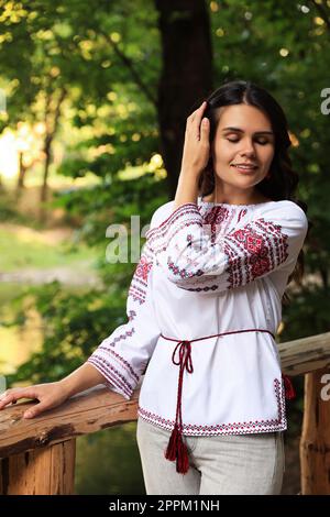 Schöne Frau, die ein besticktes Hemd neben einem Holzgeländer auf dem Land trägt. Ukrainische Nationalkleidung Stockfoto