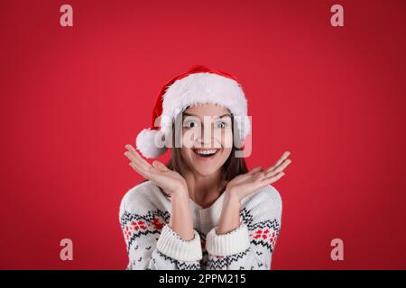 Überraschte Frau mit Weihnachtsmannmütze und Weihnachtspullover auf rotem Hintergrund Stockfoto