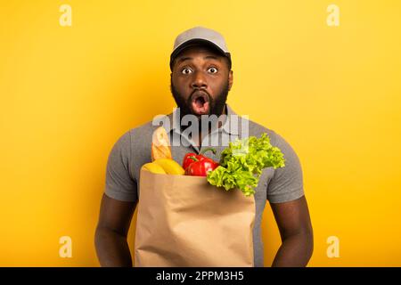 Deliveryman mit wunderten Ausdruck bereit, Beutel mit Lebensmitteln zu liefern. Gelber Hintergrund. Stockfoto