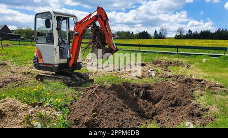 Arbeiten auf der Baustelle eines Hauses. Der Bagger gräbt Wurzeln aus. Ein kleiner Graber im Garten. Stockfoto