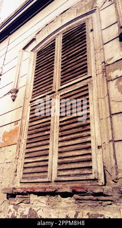 Geschlossene Fensterläden an einem hohen Fenster mit Plattbändern. Die Fassade des Gebäudes, die lange Zeit nicht repariert wurde. Der Putz blättert unter dem Fenster ab. Die Architektur der alten europäischen Stadt. Stockfoto