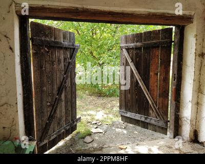 Wunderschöne alte, offene Holztür. Innenansicht des Zimmers. Kinderbettüren, Stall für Vieh oder Garage. Holzbalken und Türrahmen. Sommerliches Grün von der Tür Stockfoto