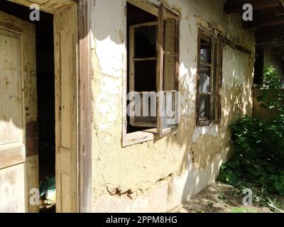 Mauer mit alten Fenstern und einer Tür in einem einbrechenden Landhaus. Der Begriff Urbanisierung und Vergessenheit des Mutterlandes. Verlassenes Haus. Zerbrochenes Glas in den Fenstern und herunterfallender Putz Stockfoto