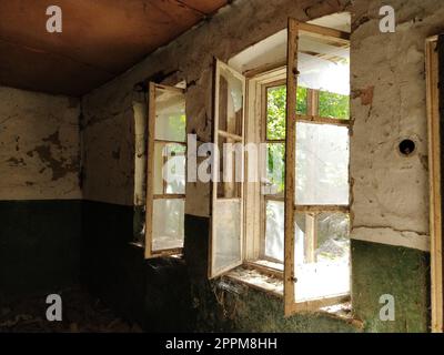 In einem verlassenen Haus. Blick auf kaputte Fenster ohne Vorhang. Grunge-Szene. Durch die alten Holzrahmen mit zerbrochenem Glas bricht warmes Sonnenlicht. Das Konzept von Veränderung, Abkehr, Nützlichkeit. Stockfoto