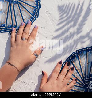Hände mit lila Nägeln und Ringen auf Tarot-Karten, die auf einer weißen Oberfläche mit Farnblatt-Schatten ausgefächert sind. Draufsicht Stockfoto
