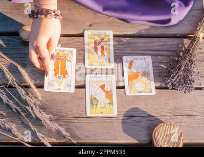 Die Hand einer Frau mit lila Nägeln legt eine von vier Tarot-Karten auf einer Holzfläche neben Stacheln und Lavendel Stockfoto