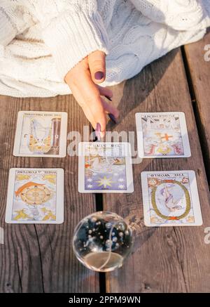 Die Hand einer Frau mit lila Nägeln zeigt auf fünf Tarot-Karten, die auf einer Holzfläche neben einer Kristallkugel verteilt sind. Vertikal Stockfoto