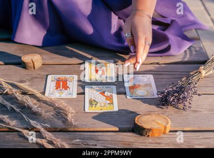 Die Hand einer Frau mit lila Nägeln zeigt auf vier Tarot-Karten, die auf einer Holzfläche neben Stacheln und Lavendel verteilt sind Stockfoto