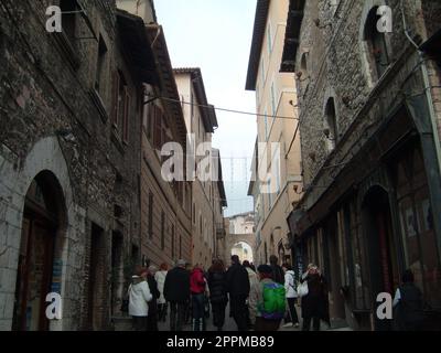 Assisi, Italien, 10. Dezember 2020. Straßen von Assisi, entlang derer eine Gruppe von Touristen zu Fuß geht. Alte Gebäude, nahe beieinander. Leute auf Ausflügen. Enge Straße Stockfoto