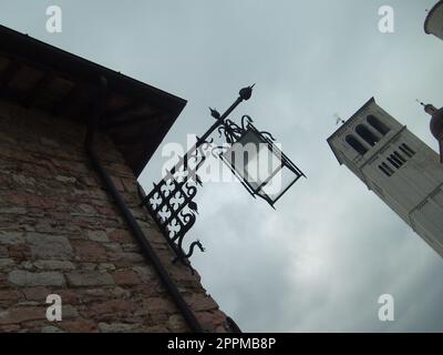 Assisi, Italien, 1. Dezember 2007 Foto aus einem Blickwinkel. Blick von unten auf einen alten italienischen Turm und eine wunderschöne Straßenlampe aus Metall. Backsteinwand eines Gebäudes mit einer pfeilförmigen Laterne. Stockfoto