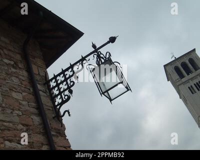 Assisi, Italien, 1. Dezember 2007 Foto aus einem Blickwinkel. Blick von unten auf einen alten italienischen Turm und eine wunderschöne Straßenlampe aus Metall. Backsteinwand eines Gebäudes mit einer pfeilförmigen Laterne Stockfoto