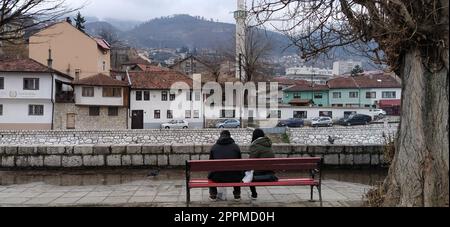 Sarajevo, Bosnien und Herzegowina - 08. März 2020. Ein junges Paar, das auf einer Holzbank unter einem Baum sitzt. Der Ufer des Flusses Milyack. Blick auf die Stadt Sarajevo, eine Moschee und ein Minarett Stockfoto