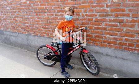 Belgrad, Serbien, 13. April 2020: Ein blonder, gutaussehender Junge von 7 Jahren trägt eine blaue schützende chirurgische Maske auf seinem Gesicht. Das Kind fährt Fahrrad, hält an einer Mauer und schaut in die Kamera Stockfoto