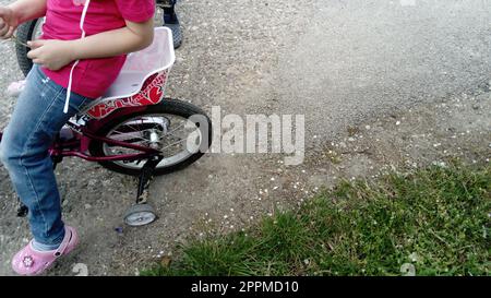 Belgrad, Serbien, 13. April 2020: Hinterrad eines Kinderfahrrads mit kleinem Zusatzrad. Teilweise sichtbar sind der Körper und das Bein eines Mädchens, das auf einem Fahrradsitz sitzt. Asphalt und grünes Gras Stockfoto