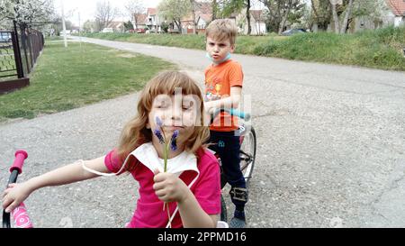 Belgrad, Serbien, 13. April 2020: Kinder in chirurgischen Schutzmasken fahren Fahrräder. Die Kinder nahmen ihre Masken ab. Das Mädchen schnüffelt an einer lila Blume und lächelt, und der Junge runzelt die Stirn und sagt Stockfoto